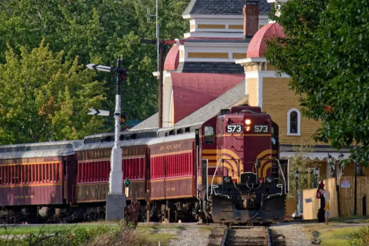 Vintage red train arriving at a picturesque station with lush green trees in the background.