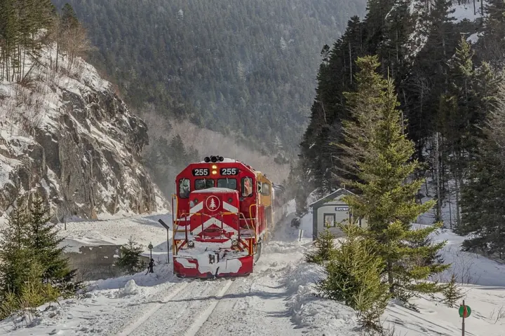 Red train travels through snowy mountain landscape with pine trees and rocky cliffs.