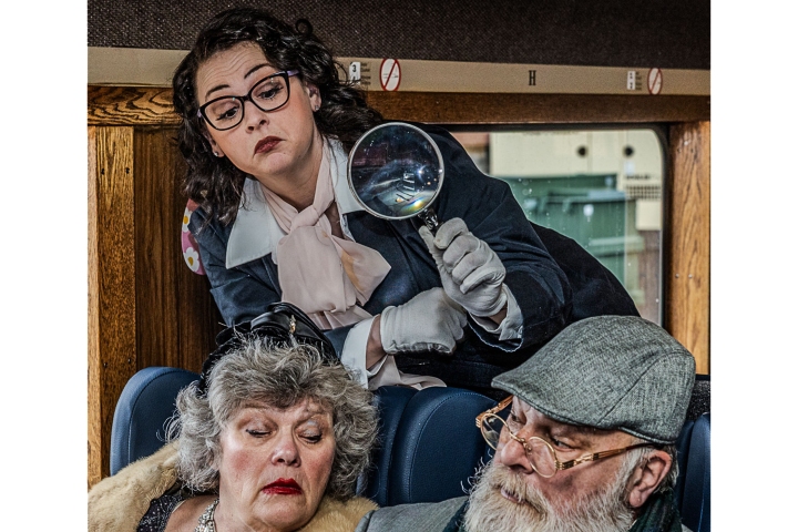 Person in vintage clothes examines passengers with a magnifying glass on a train.