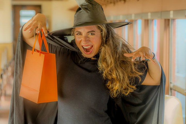Woman in witch costume holding an orange bag, standing indoors.