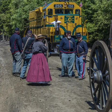 People in historical clothing near a yellow train and wooden wagon wheel on a dirt road.