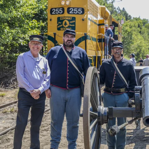 Three men in uniform stand beside a cannon in front of a yellow train.
