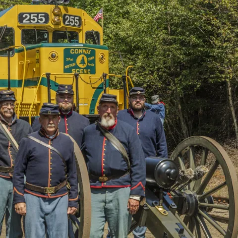 Five men in Civil War uniforms stand by a cannon in front of a yellow train labeled Conway Scenic.