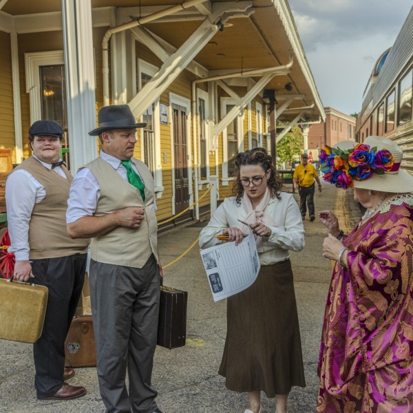 Four people in vintage attire at a train station with luggage and a train in the background.