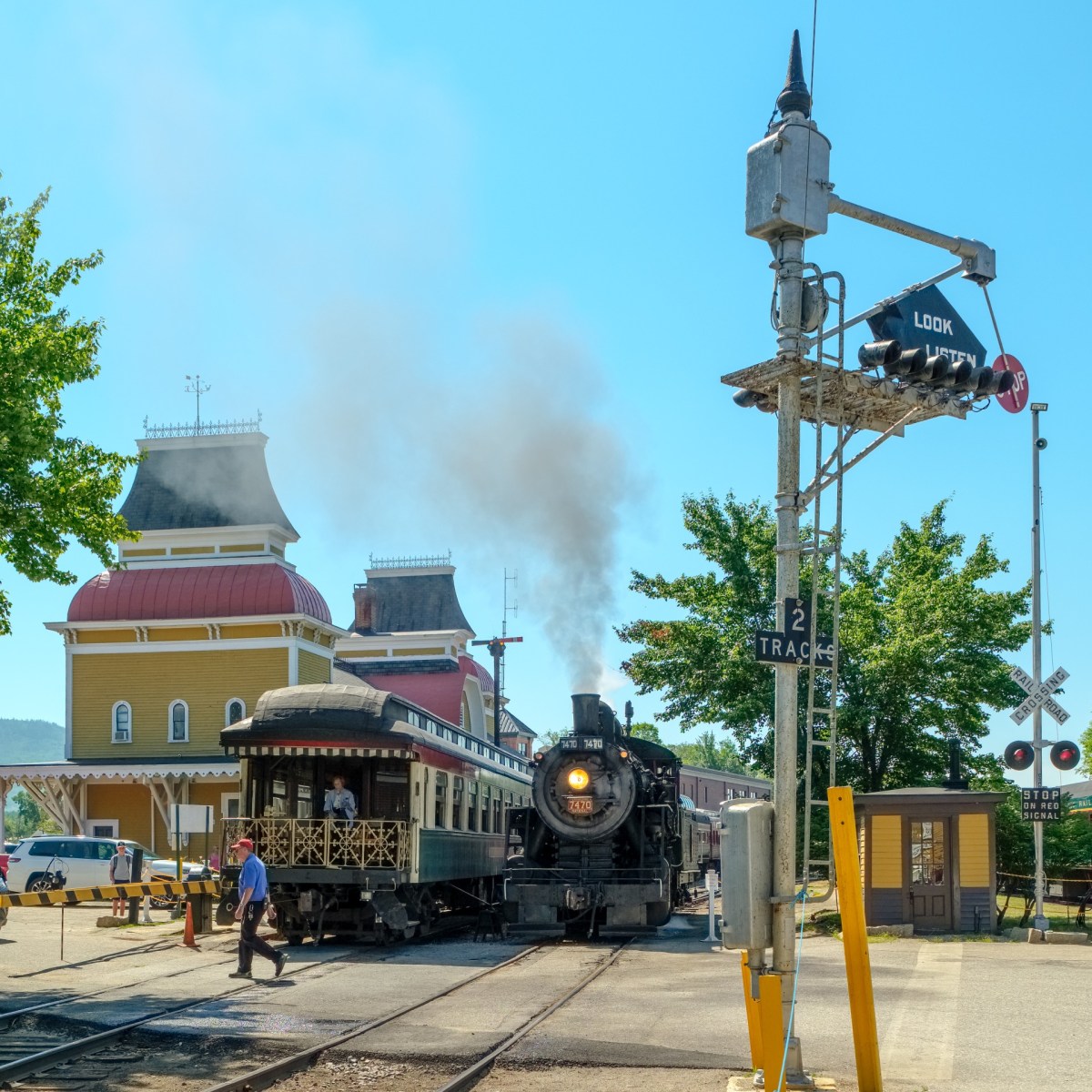 Steam train near a vintage station with people, trees, and railroad crossing signs under a clear blue sky.