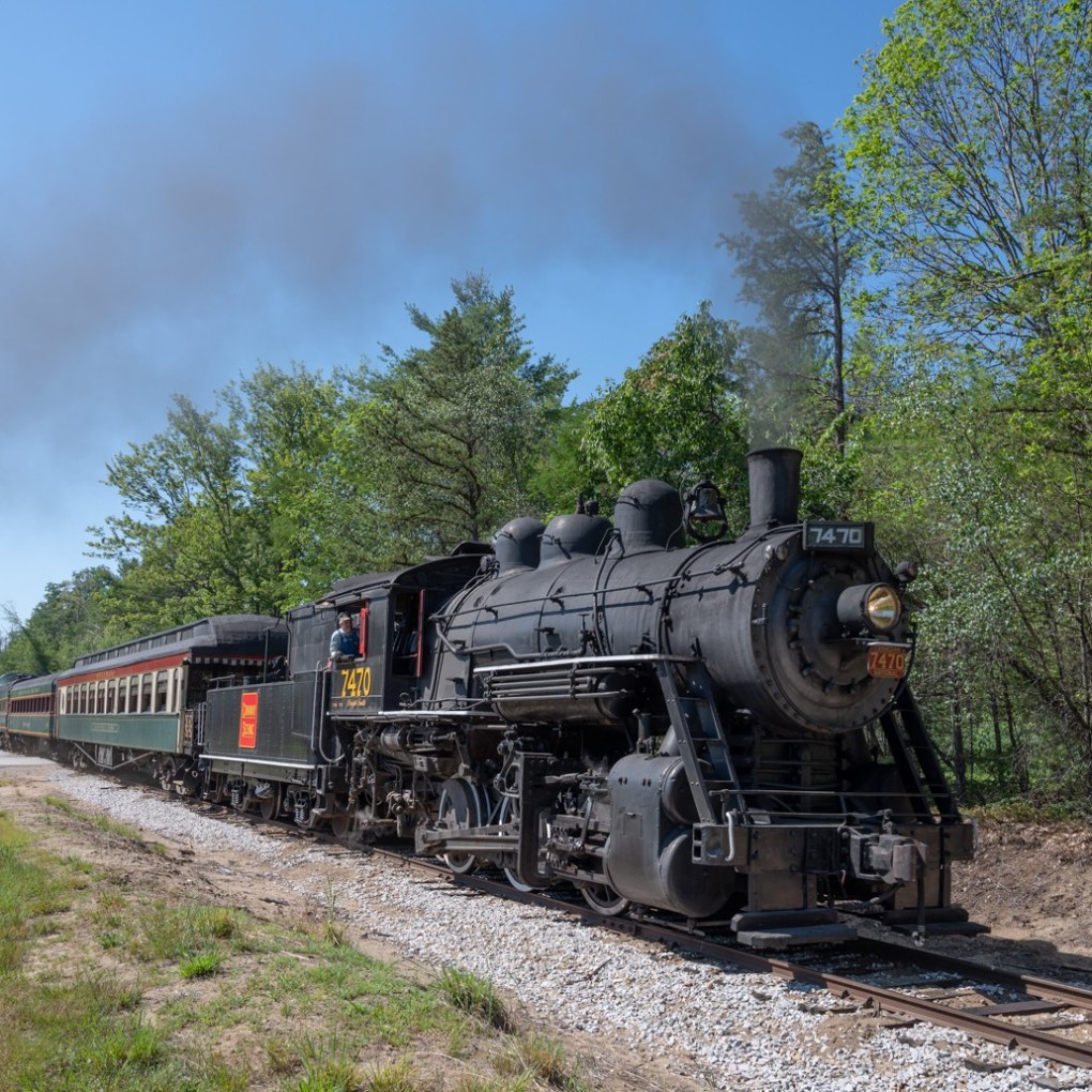 Steam locomotive releasing smoke while traveling through a wooded area on a clear day.