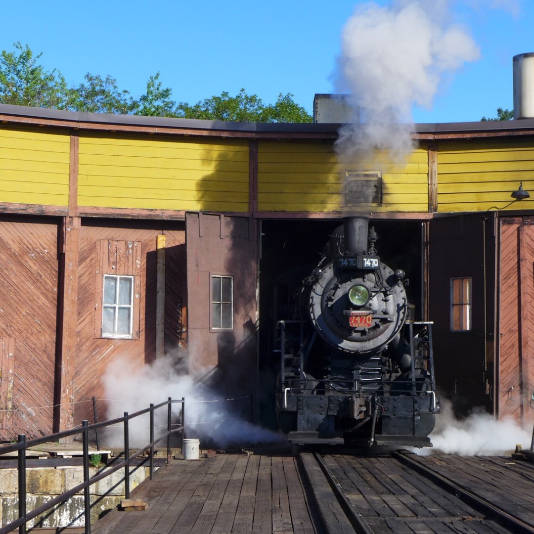 Steam locomotive exiting wooden roundhouse with yellow trim and steam rising.