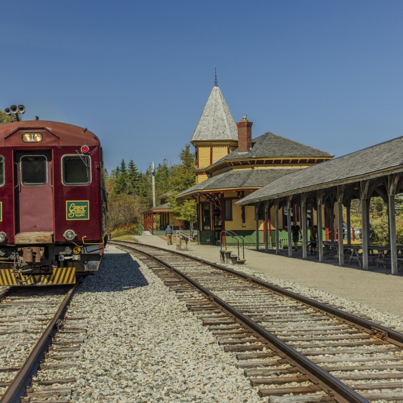 Red train at small rural station with canopy, surrounded by trees under clear sky.