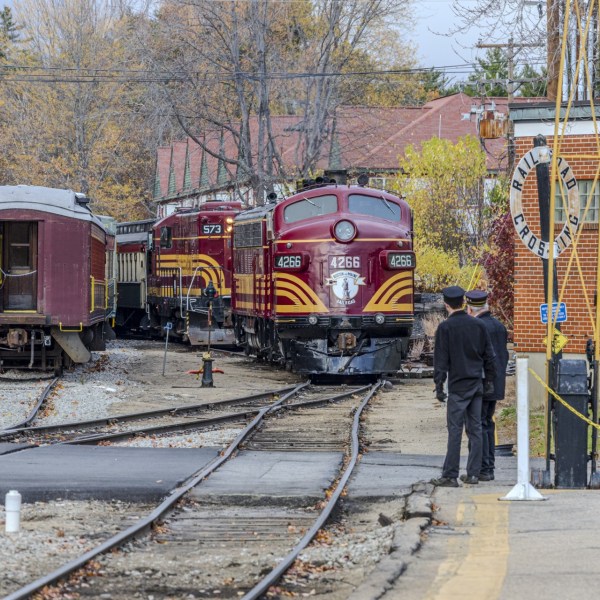 Vintage train with maroon locomotives at a railroad crossing, surrounded by people and autumn trees.