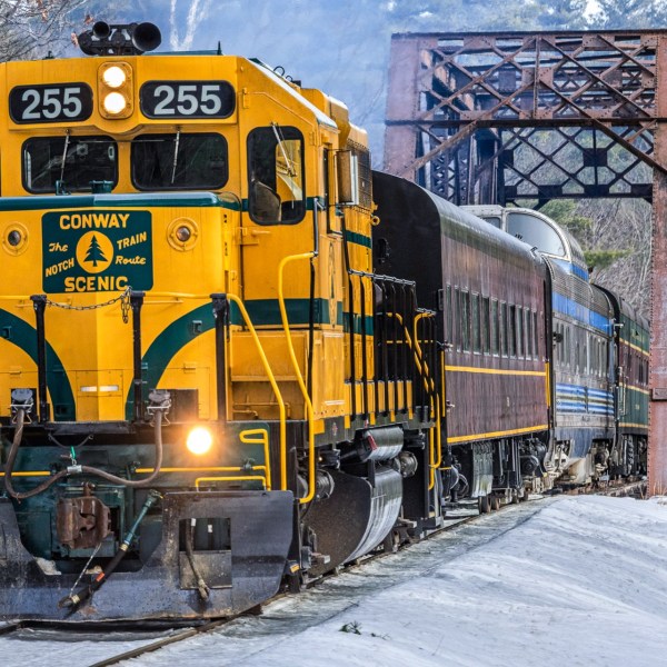 Yellow scenic train crossing a metal bridge in a snowy forest landscape.