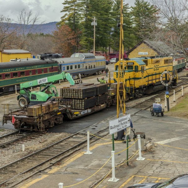 Railroad yard with a green excavator on a train car, alongside colorful trains and buildings.