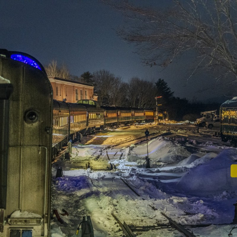 Snowy train tracks with vintage trains at dusk, surrounded by trees and a building.