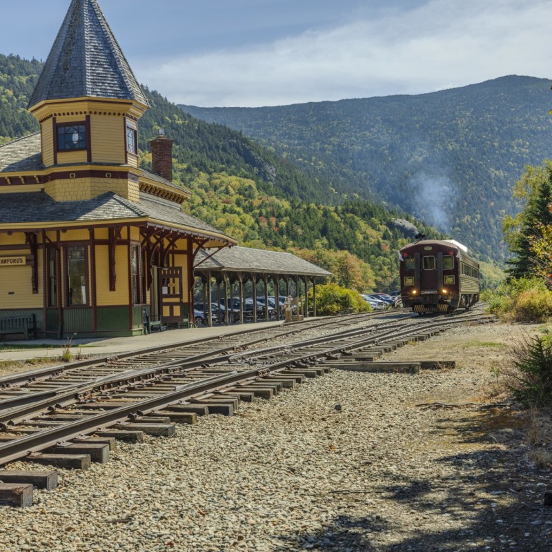 Train approaching a vintage station in a scenic mountain setting on a clear day.