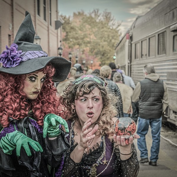 Two people in witch costumes pose on a train platform with a crowd in the background.
