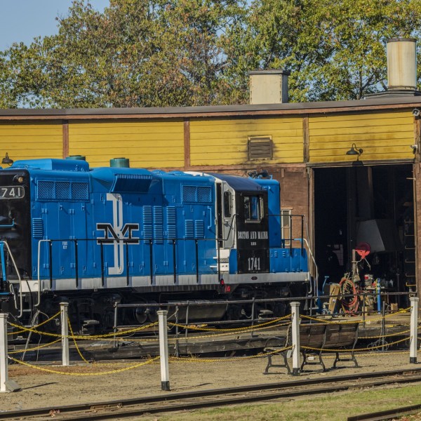Blue diesel locomotive in front of yellow maintenance shed with trees in background.