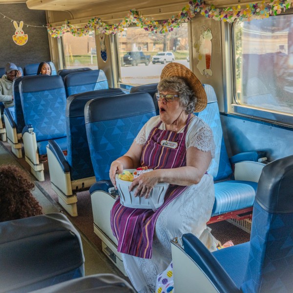 Woman in hat and apron sitting on a train seat, holding a box, with passengers in the background.