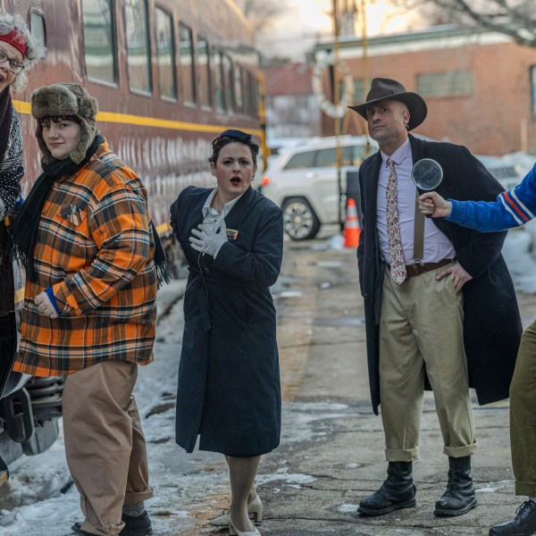 Five people in vintage attire outside a train, one holding a magnifying glass, snowy ground.