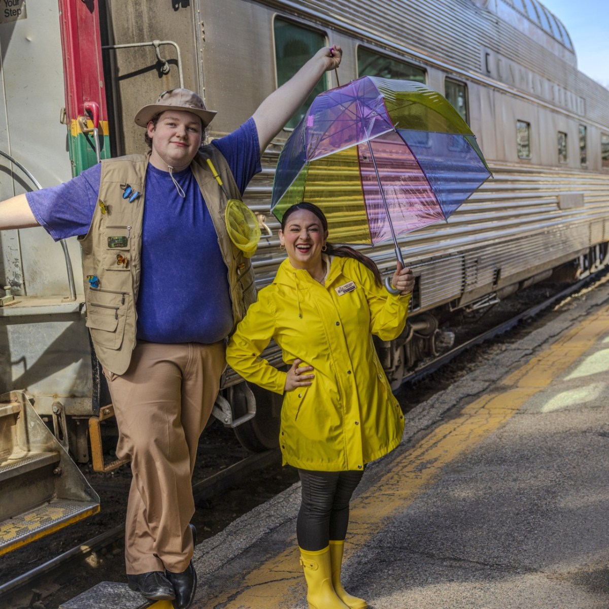Two people in rain gear posing happily next to a train on a station platform.