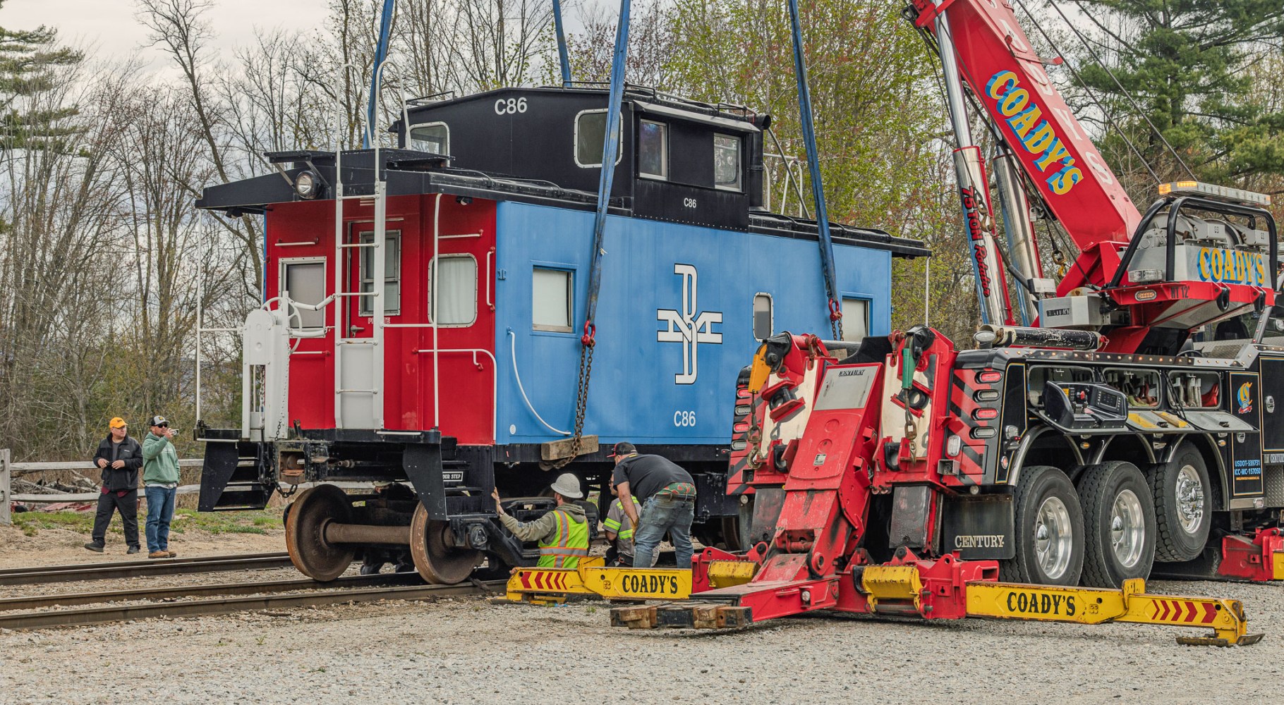 Red and blue train caboose being lifted by large crane vehicle.