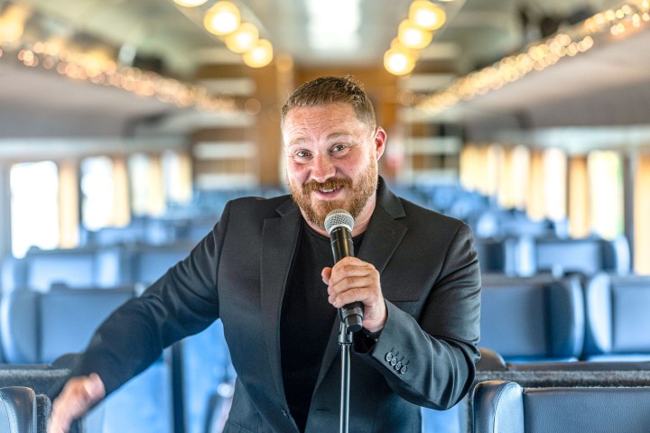 Man in a suit holding a microphone, standing in a train aisle.