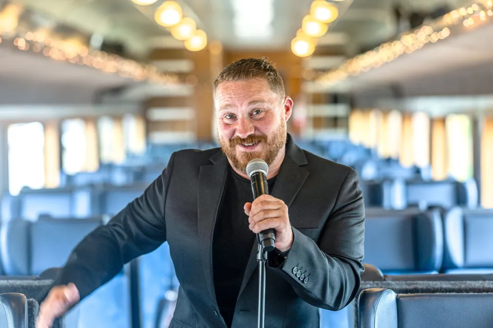 Man in a suit holding a microphone, standing in a train aisle.
