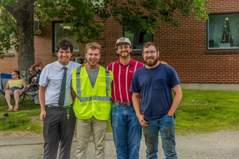 Four men smiling outdoors, one wearing a safety vest, in front of a brick building.