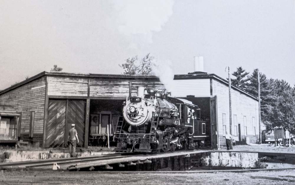 Black and white photo of a steam locomotive exiting a wooden building.
