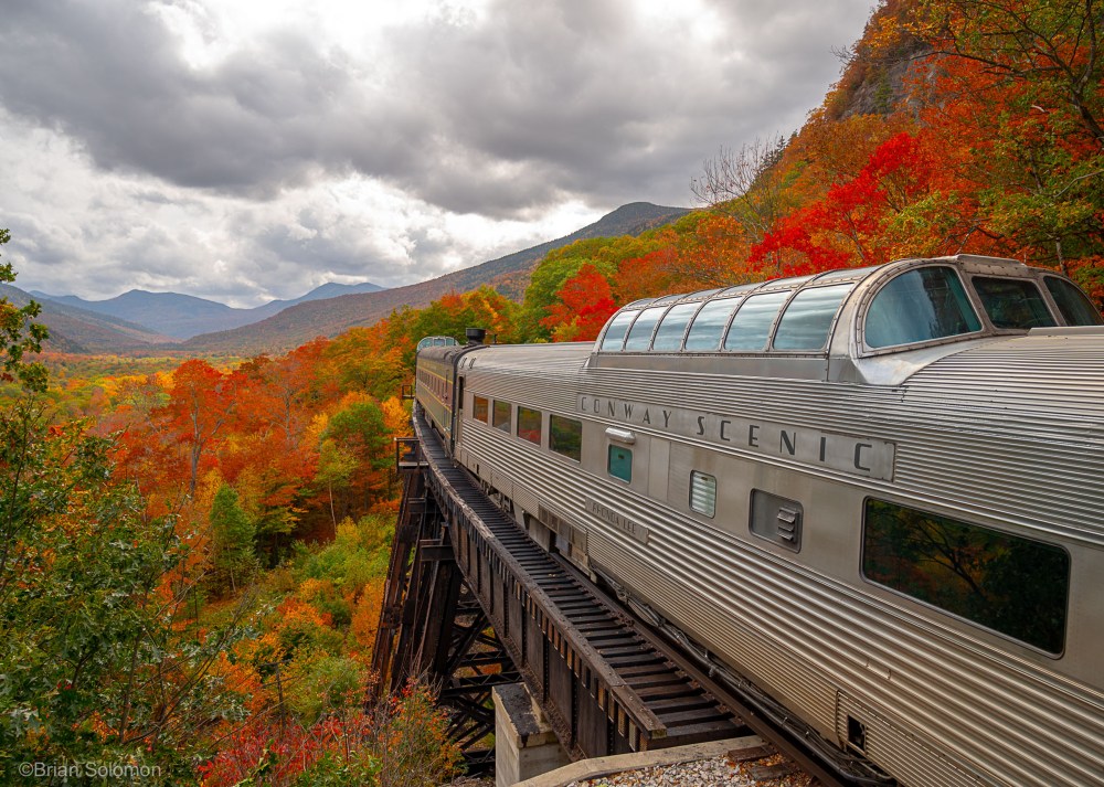 Train on a bridge amidst vibrant autumn foliage in a mountainous landscape under cloudy skies.