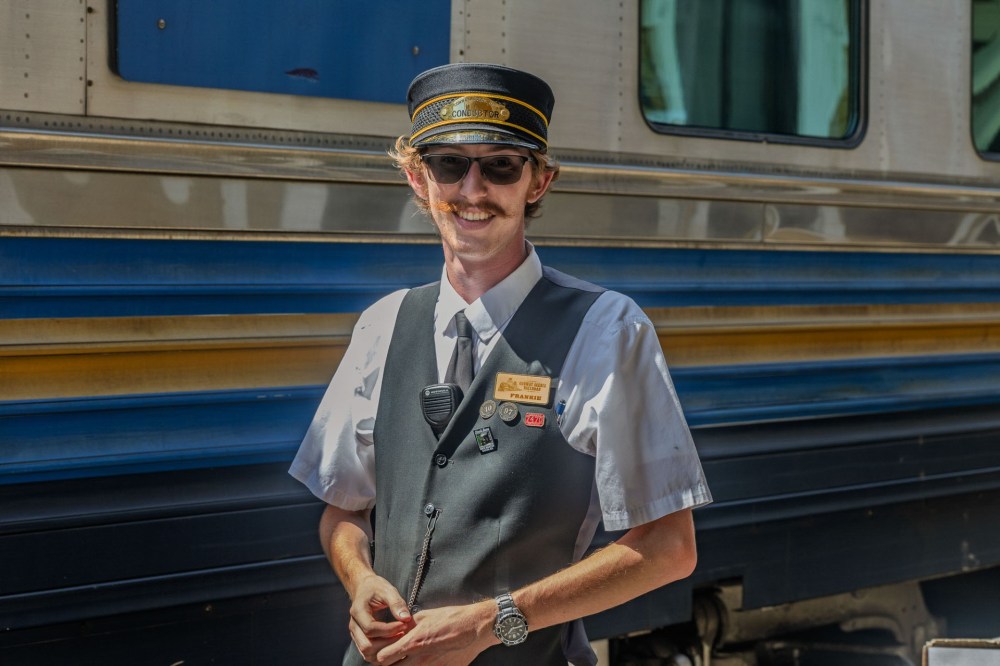Train conductor in uniform with sunglasses stands near a train.