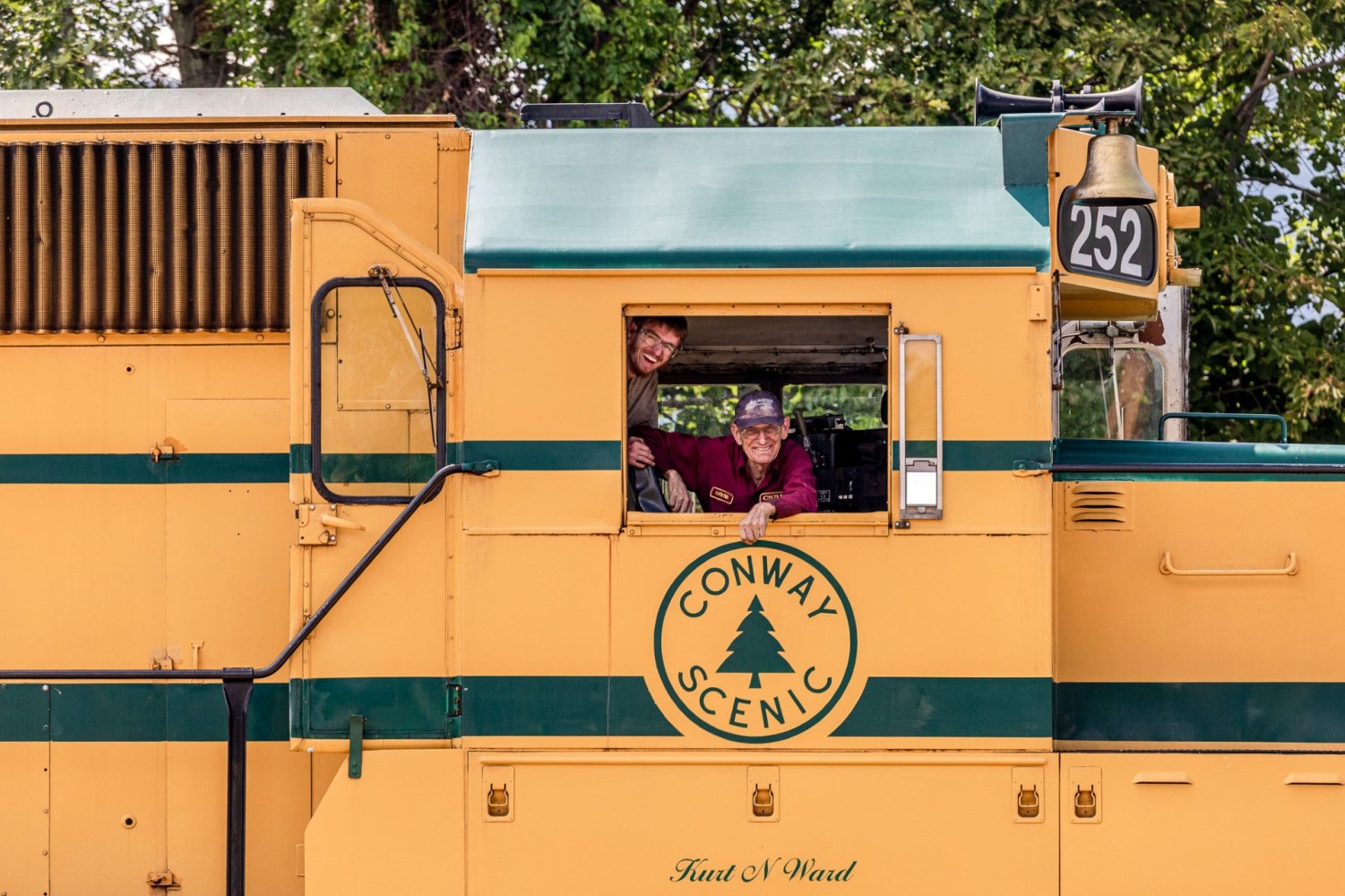 Two people smiling from a yellow train cab with 'Conway Scenic' logo.